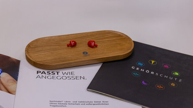 Auf 2 Broschüren zum Thema Gehörschutz, liegen zwei kleine Hörgerätsohrstücke in roter Farbe auf einer hölzernen Unterlage.