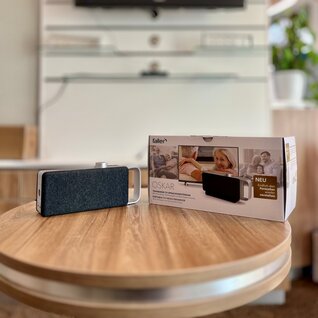 Tragbarer TV-Sprachverstärker in schwarz mit dem Produktnamen OSKAR, welcher sich links auf einem Tisch neben dessen Verpackungsbox im Laden von Hörgeräte Jens Steudler befindet. 