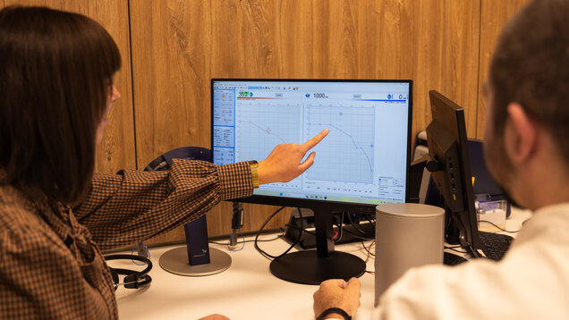 Eine Frau zeigt auf einen PC Bildschirm auf dem ein Audiogramm abgebildet ist. Neben Ihr sitzt ein Mann, dem Sie die Grafik zeigt.